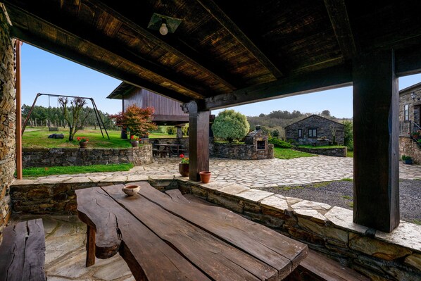 A rustic outdoor patio with a wooden table and benches, stone walls, and a view of a garden and buildings in the distance.