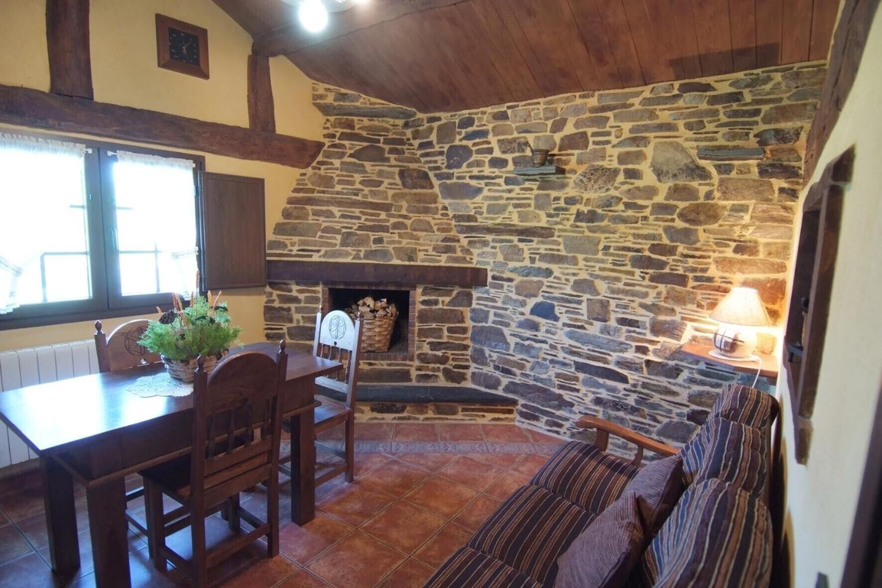 Chimenea de piedra con leña encendida, mesa de comedor de madera con sillas y una zona de estar con cojines.