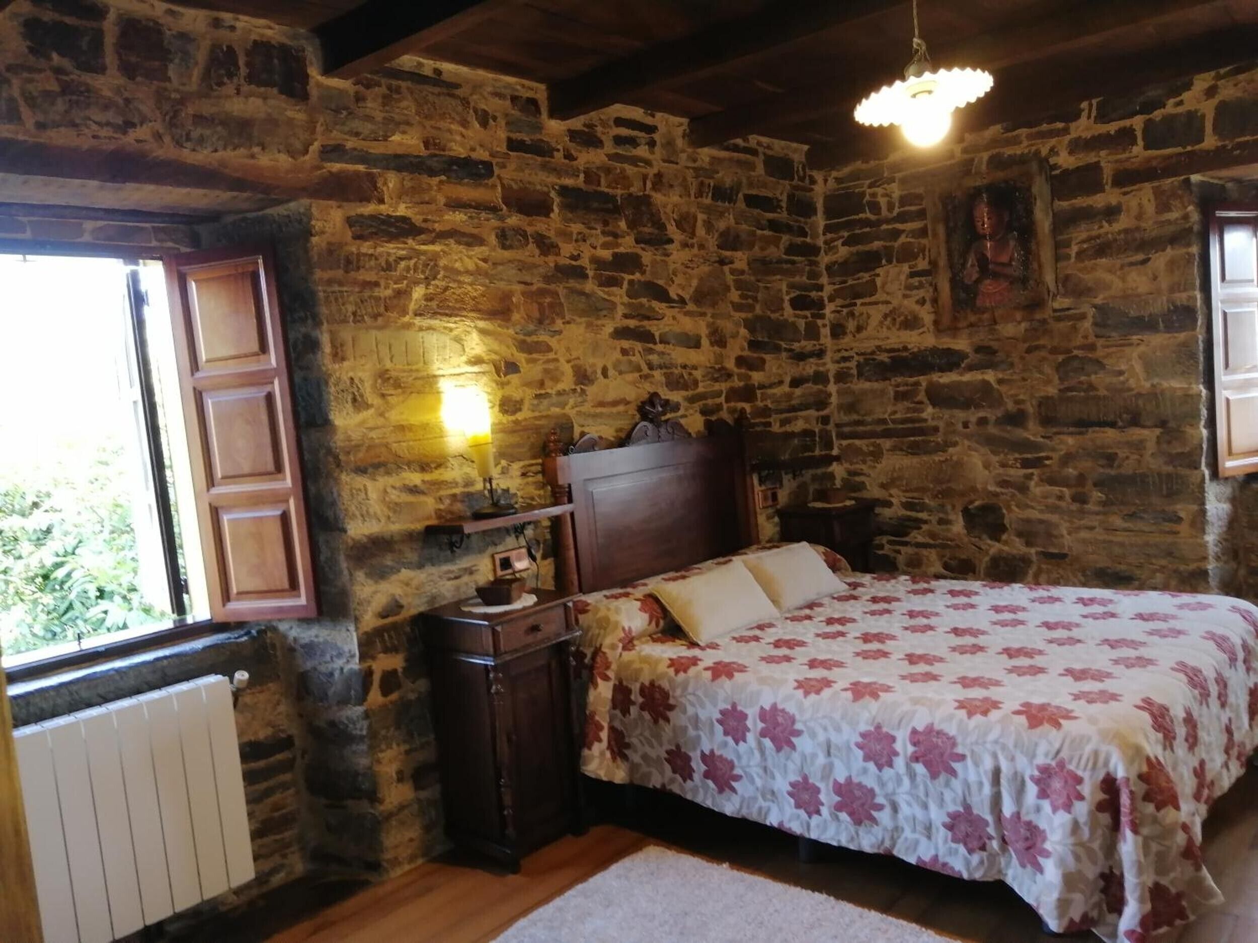 Een slaapkamer met een stenen muur, een houten bed, een nachtkastje, een lamp en een raam met houten luik.