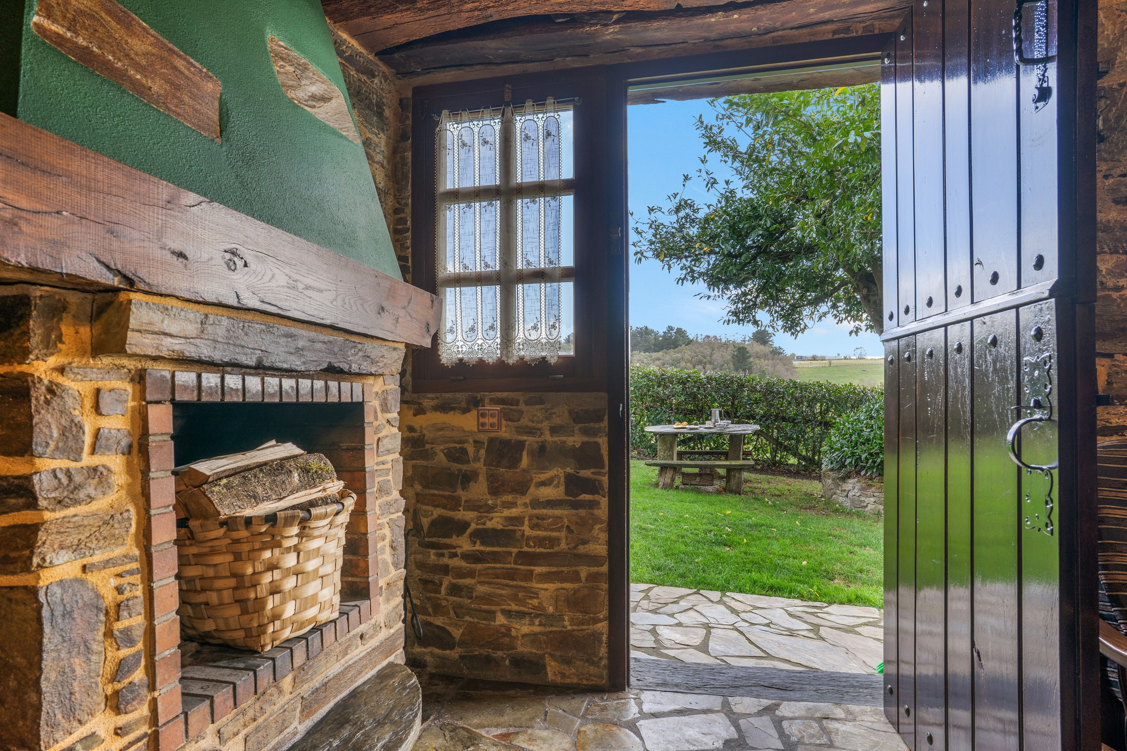 Una chimenea de piedra rústica con una cesta tejida, techo de madera y vista a un jardín a través de una puerta de madera.