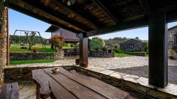 A rustic outdoor patio with a wooden table and benches, stone walls, and a view of a garden and buildings in the distance.