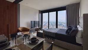 Quarto de hotel moderno com uma cama grande, um sofá, uma televisão e um espaço de jantar com mesa posta para dois.