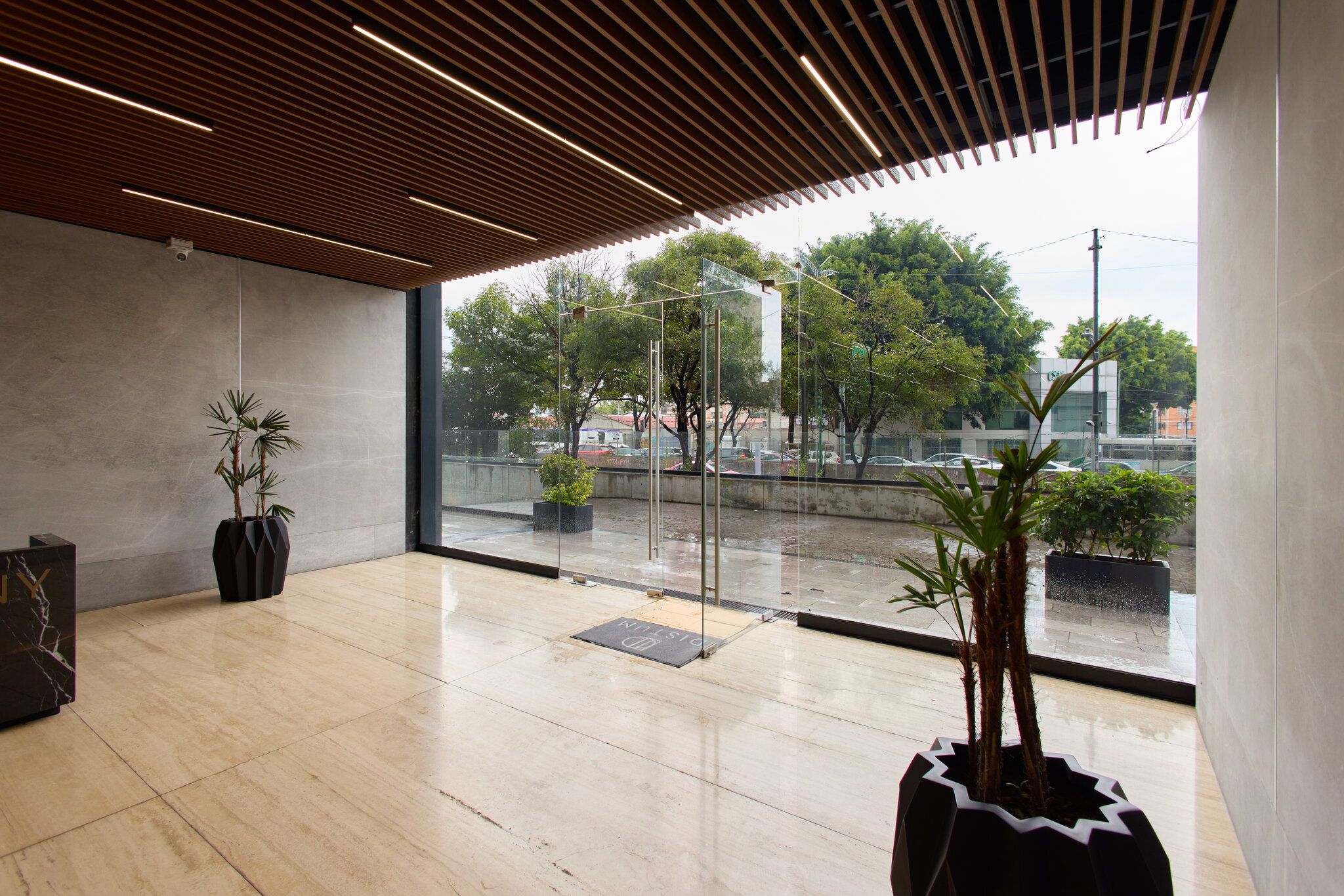 A spacious indoor area with a modern design, featuring a wooden ceiling, large windows, and potted plants.