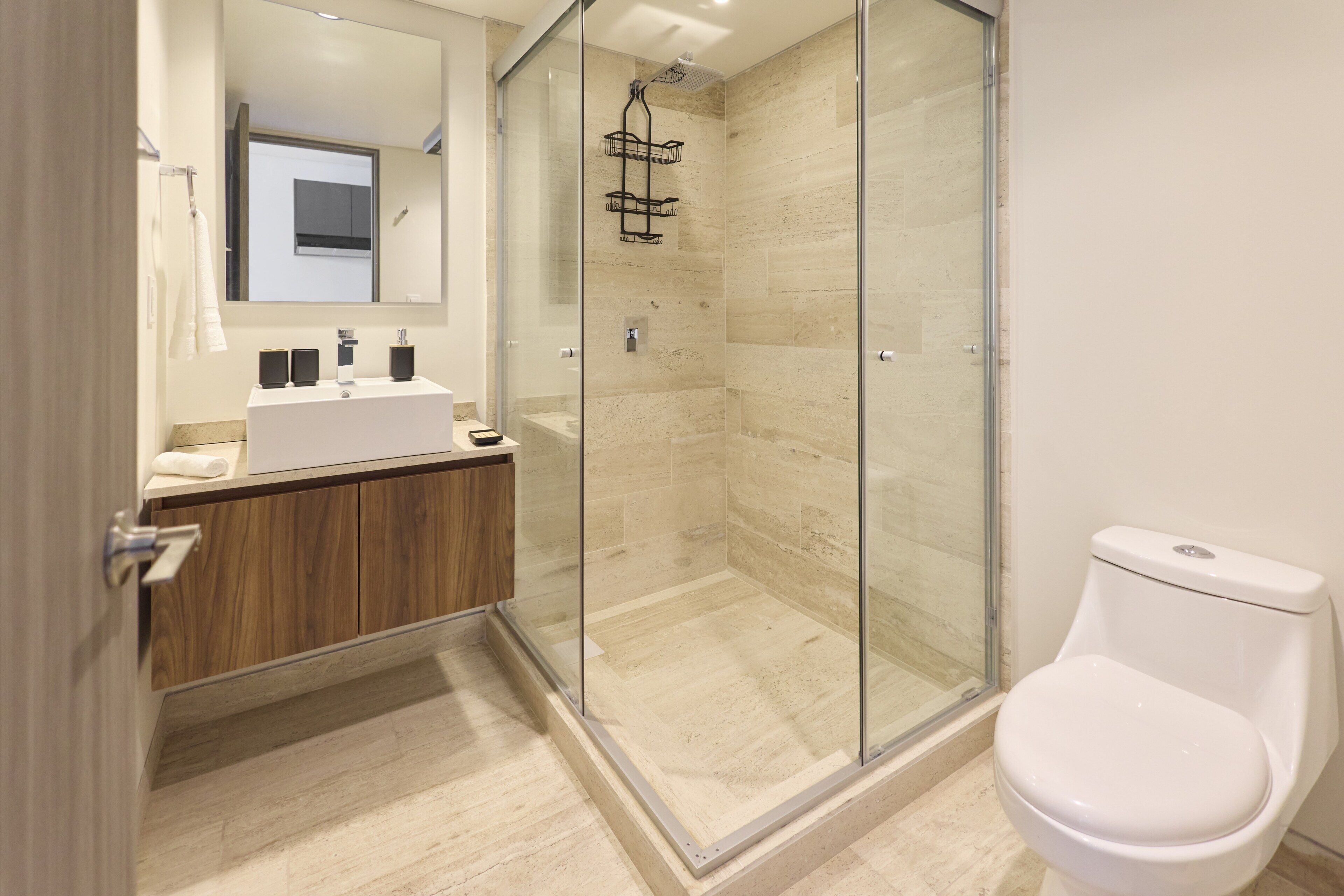 Un baño moderno con ducha envuelta en vidrio, inodoro blanco y tocador de madera con espejo.