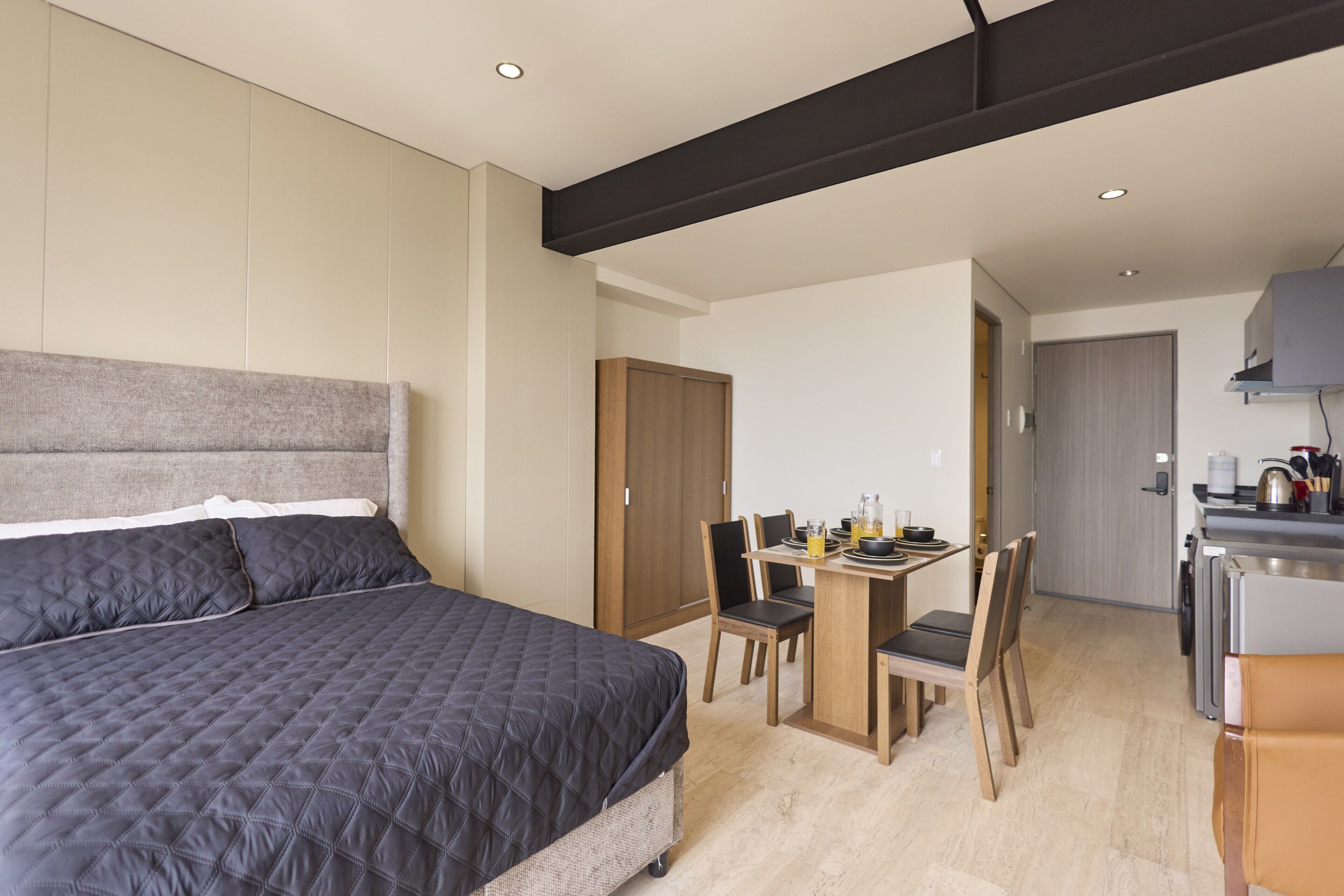A modern bedroom with a large bed, a wooden wardrobe, and a kitchenette area with a dining table and chairs.