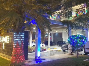 Un bâtiment doté d’une entrée couverte, d’un espace de stationnement avec des voitures et d’un arbre décoré de guirlandes lumineuses.
