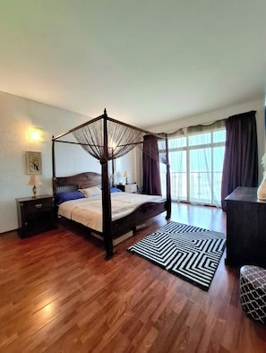 Un lit à baldaquin, une table de chevet avec une lampe, un tapis noir et blanc, et une vue sur l’extérieur grâce à de grandes fenêtres.