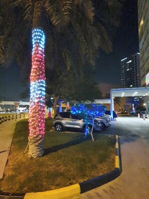 A parking area with a tree wrapped in lights, a car, and a building in the background. - Amazing 2-bedrooms flat.Full lagoon,sea view high floor. Nicely furnished,AC. (Muharraq)