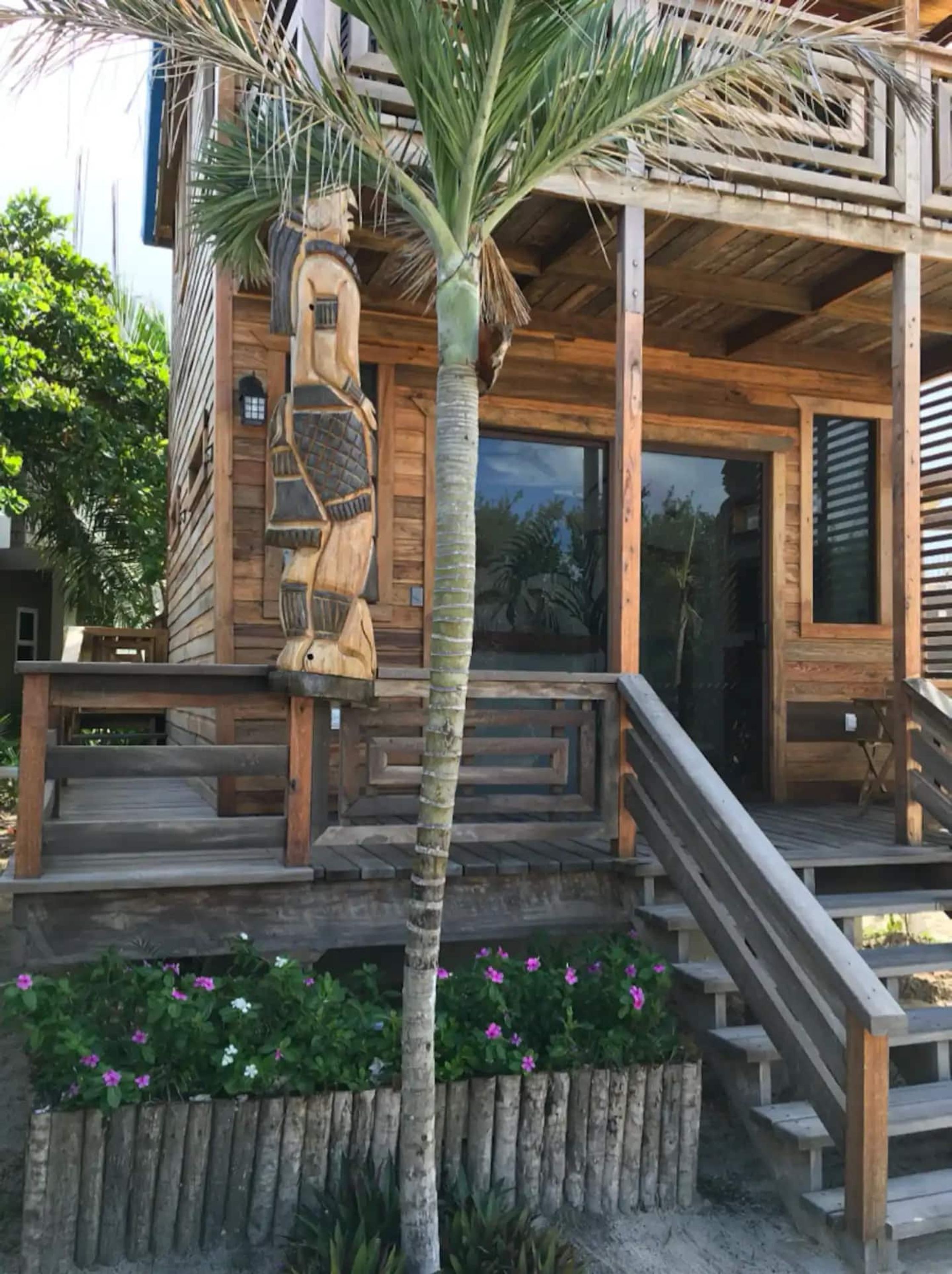 A wooden building with a staircase leading to a covered entrance, surrounded by a wooden fence and flowering plants.