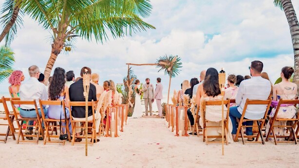 Una ceremonia de boda en la playa con invitados sentados en sillas de madera frente a un pasillo central.