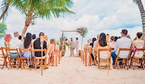 Una ceremonia de boda en la playa con invitados sentados en sillas de madera frente a un pasillo central.