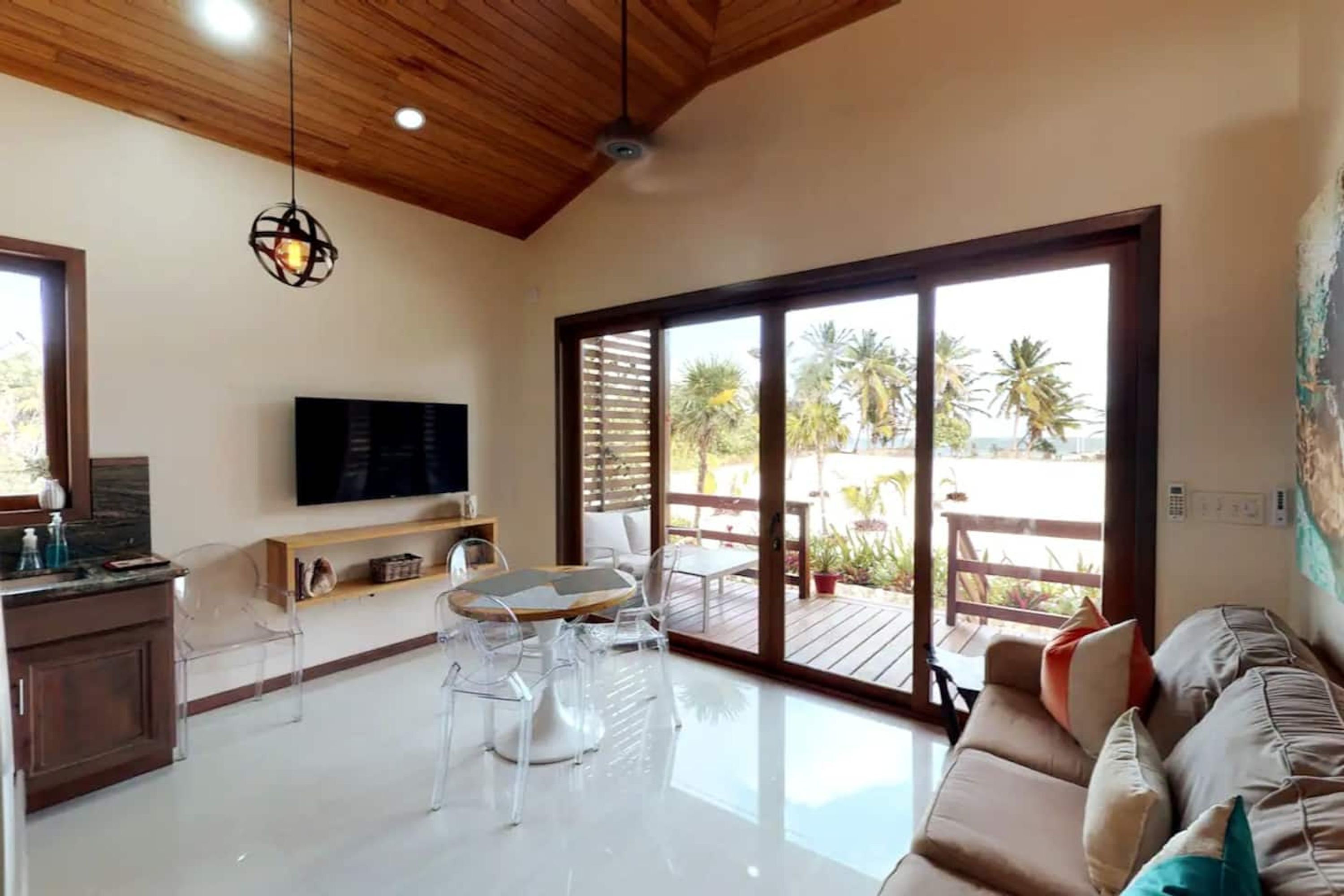 Un amplio salón con un gran ventanal corredera que da a una terraza exterior, un televisor de pantalla plana montado en la pared y un techo de madera.