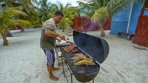 Una persona preparando comida a la parrilla al aire libre, con una barbacoa en primer plano y palmeras al fondo.