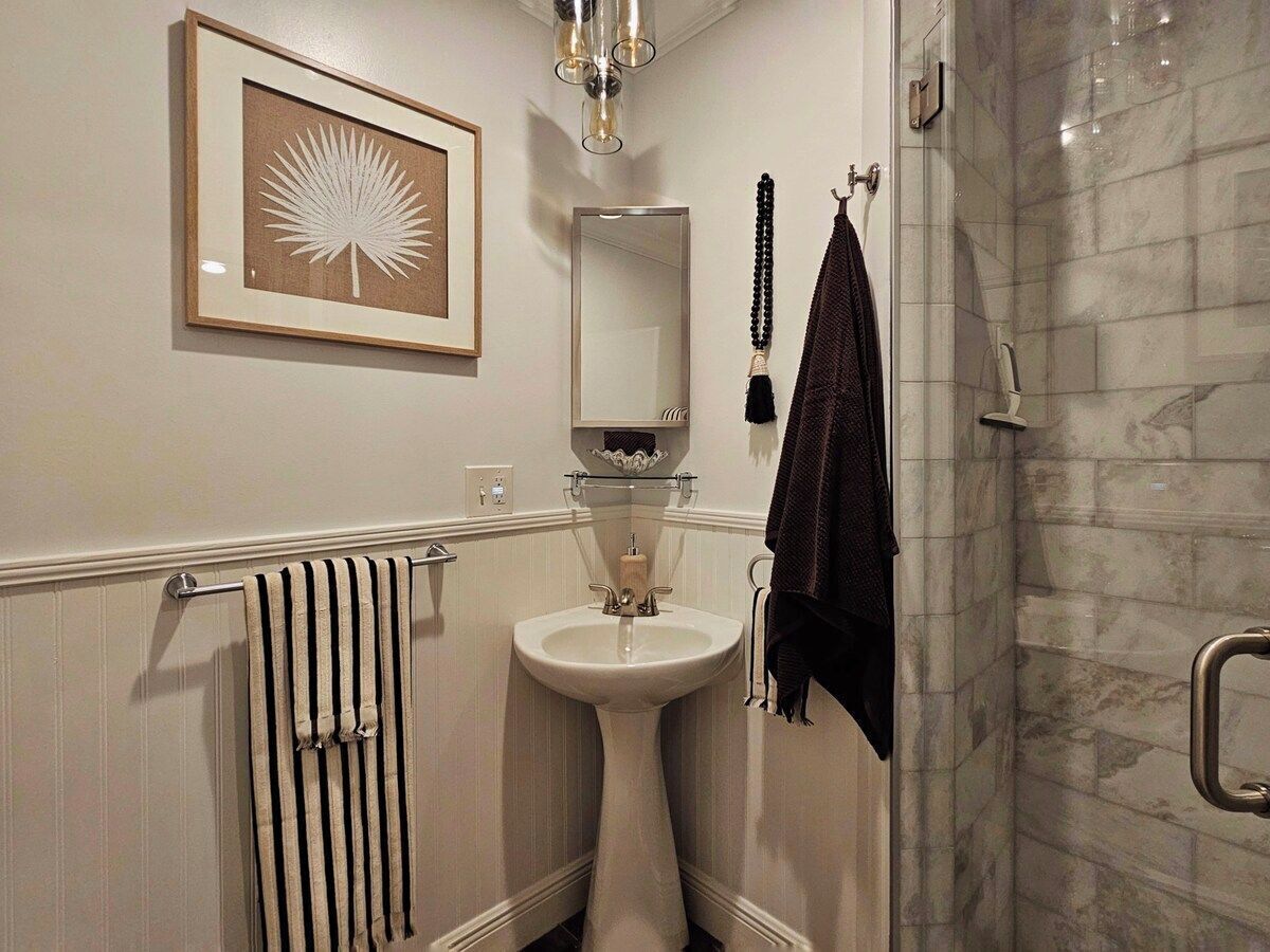 Une salle de bain avec un lavabo sur pied, une œuvre d’art encadrée au-dessus, un porte-serviettes avec une serviette foncée et une cabine de douche en verre.