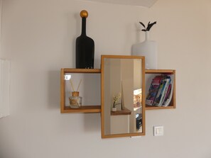 A wall-mounted shelf with a black bottle, a white vase with dried plants, and a mirror reflecting a room with a piano.