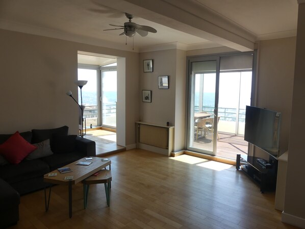Een ruime woonkamer met een plafondventilator, een bank met kussens, een salontafel, een televisie en een grote schuifpui die uitkomt op een balkon.