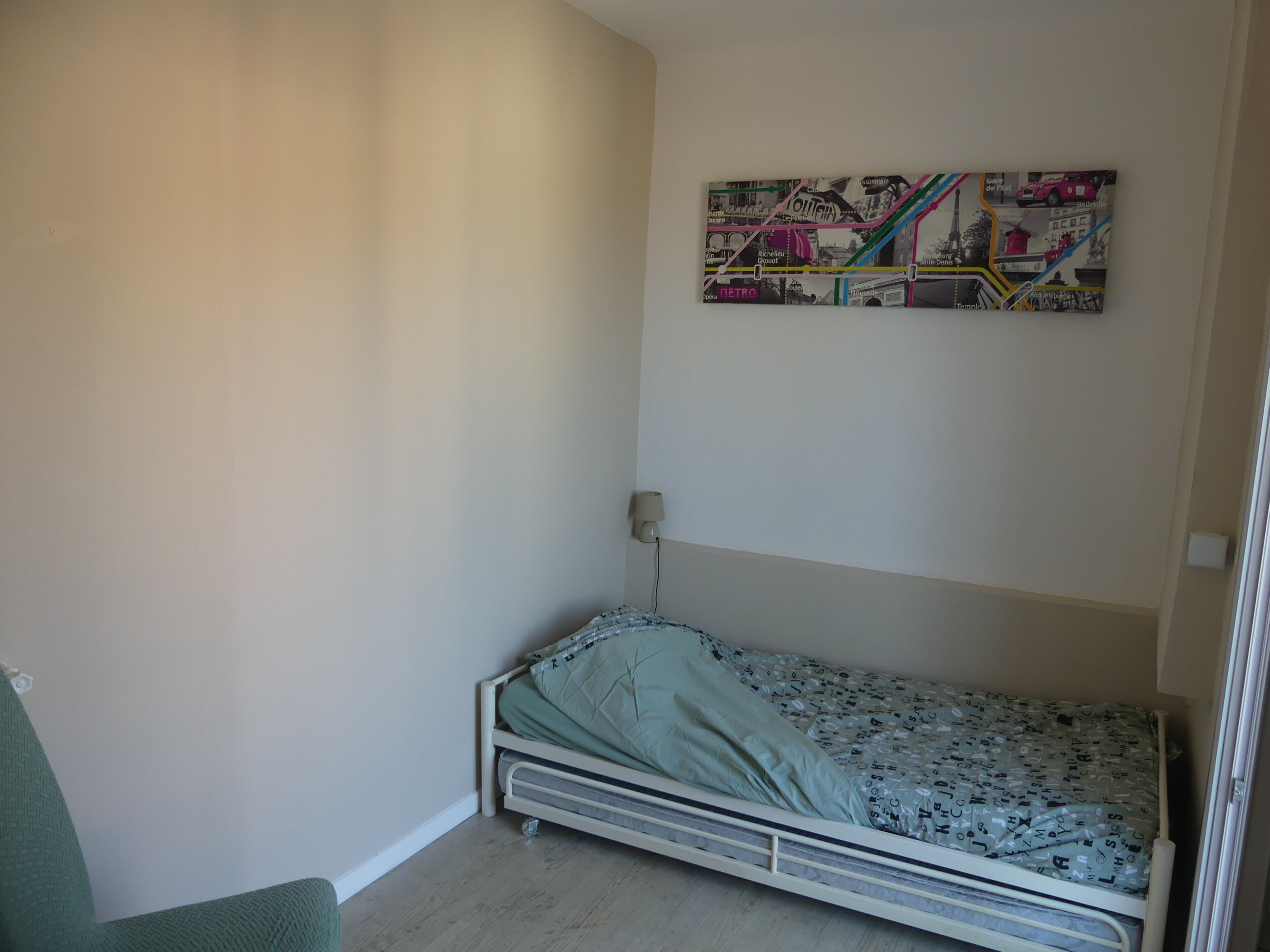 Une petite chambre avec un lit simple, un mur uni et une œuvre d’art représentant une carte aux couleurs vives.