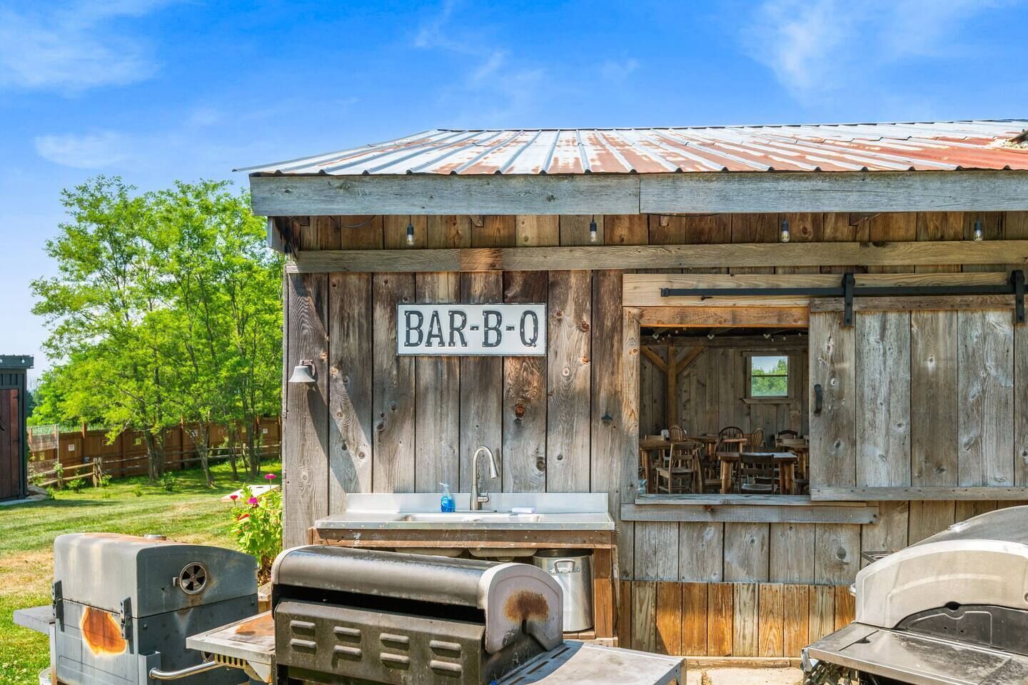 Une structure rustique en bois, avec une enseigne indiquant « BAR-B-Q » et un espace barbecue équipé d’un évier et de chaises.