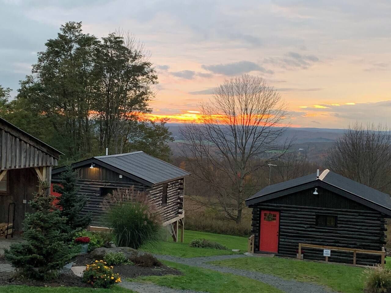 A rustic cabin with a red door, surrounded by greenery and a view of the sunset.