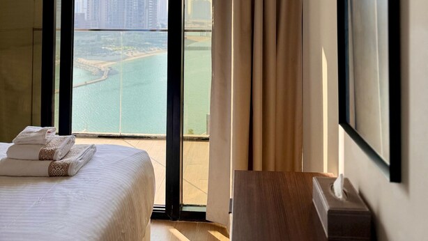 Ein Hotelzimmer mit Bett, Nachttisch mit Taschentuchbox, großem Fenster mit Blick auf Stadt und Wasser sowie einem Vorhang.