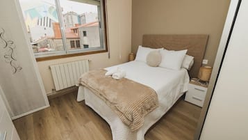 Een net opgemaakt bed met een beige deken en kussens, een wit hoofdbord, en een raam met uitzicht op gebouwen.