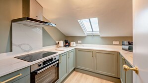 Een compacte keuken met een ingebouwde oven, een kookplaat en een magnetron. Boven de aanrecht is een dakraam en de kastjes zijn lichtgroen met gouden handgrepen.