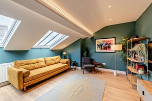 Ein modernes Wohnzimmer mit hohen Decken, Oberlichtern, einem Sofa, einem Sessel, einer Stehlampe, einem Bücherregal und einem gerahmten Bild an der Wand.