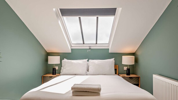 Een slaapkamer met dakraam, een bed met witte lakens, twee nachtkastjes met lampen en een radiator.