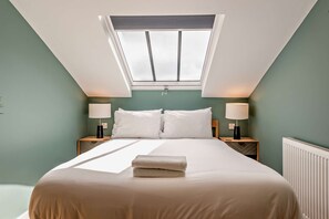 Un dormitorio con tragaluz, una cama con ropa de cama blanca, dos mesitas de noche con lámparas y un radiador.