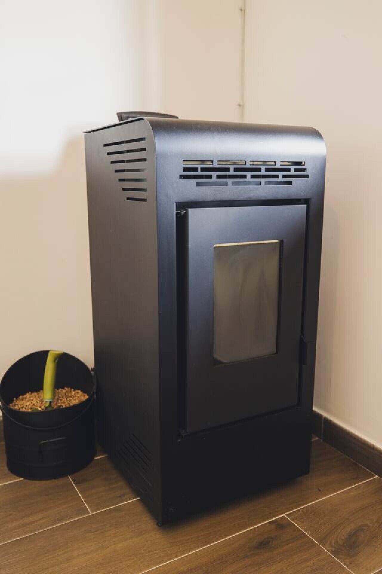 A black wood stove with a glass door, positioned next to a black trash bin on a wooden floor.