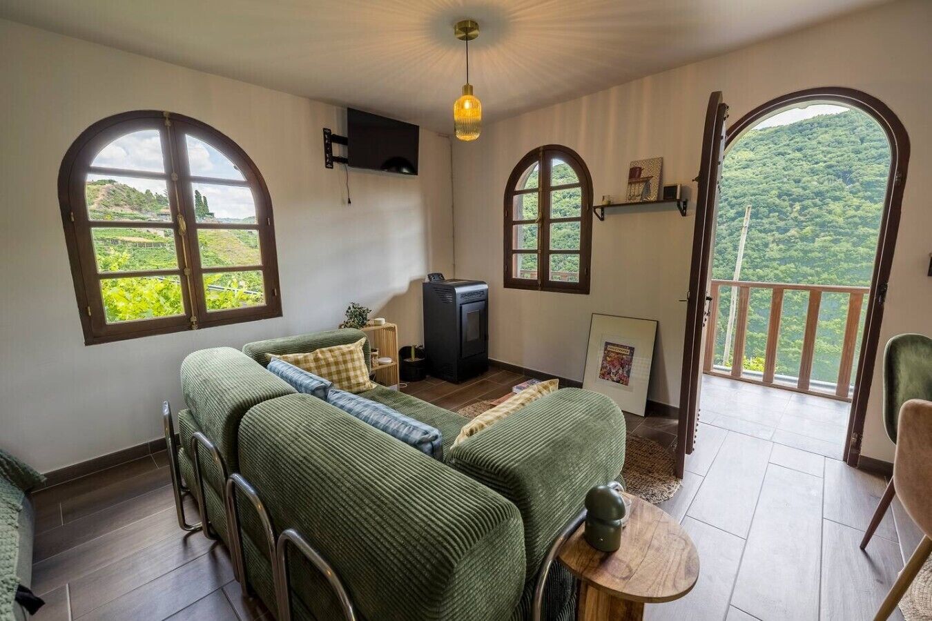A room with a sofa, a television, and a balcony with a view of greenery.