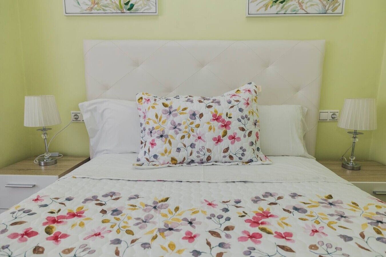 Una cama con colcha y almohadas estampadas con motivos florales, dos lámparas de noche y una pared con obras de arte enmarcadas.