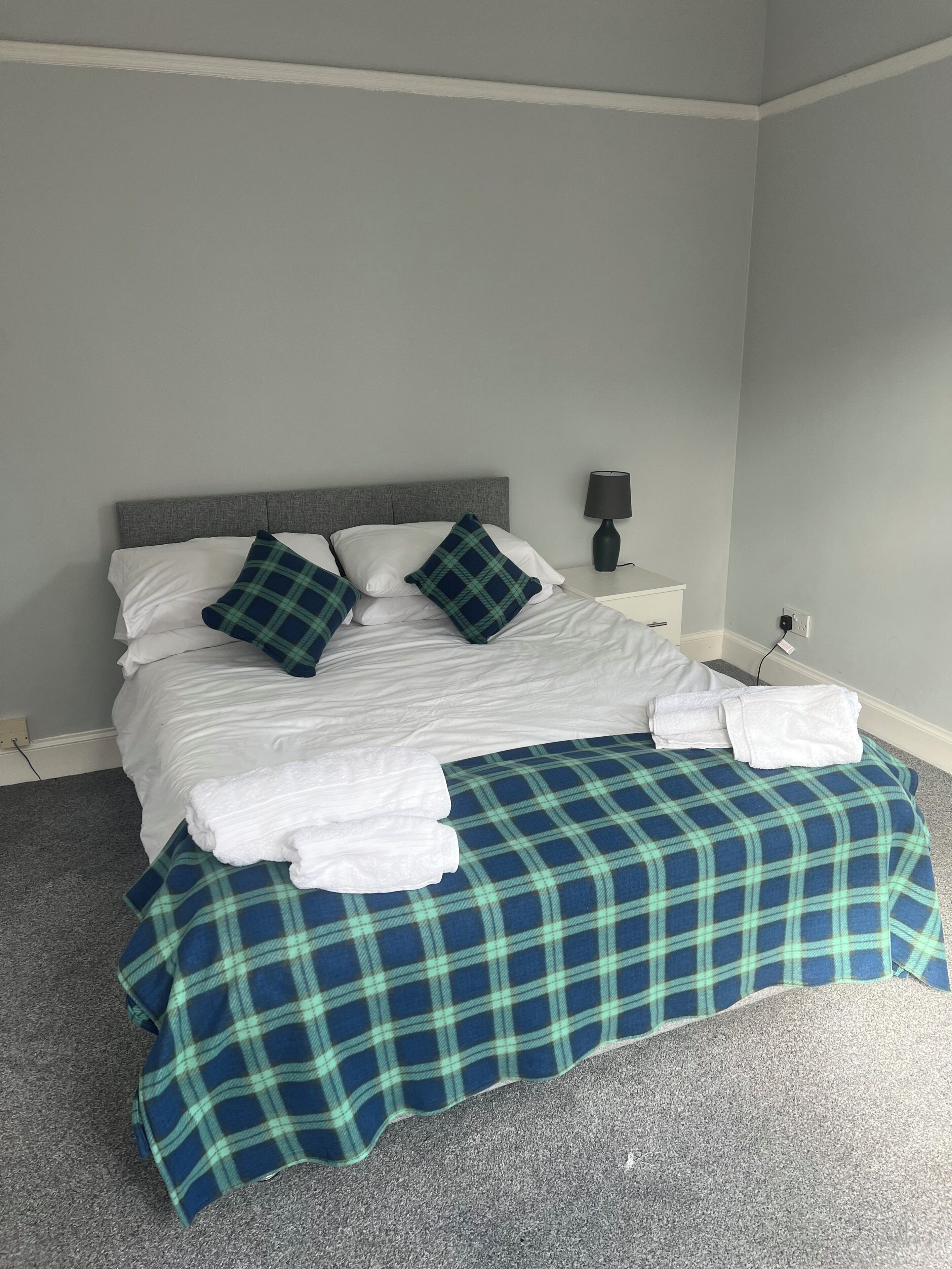 Un lit bien fait, avec du linge de lit blanc, une couvre-lit à carreaux bleus et verts, et des coussins assortis. Une lampe de chevet est posée sur une table de chevet blanche.