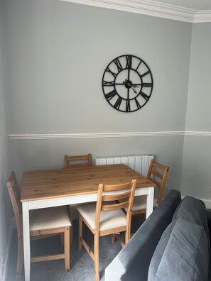 A dining area with a wooden table and chairs, a wall-mounted clock, and a radiator. -  Our Salad Days Lochgilphead in Lochgilphead town offers a spacious holiday home (Lochgilphead)