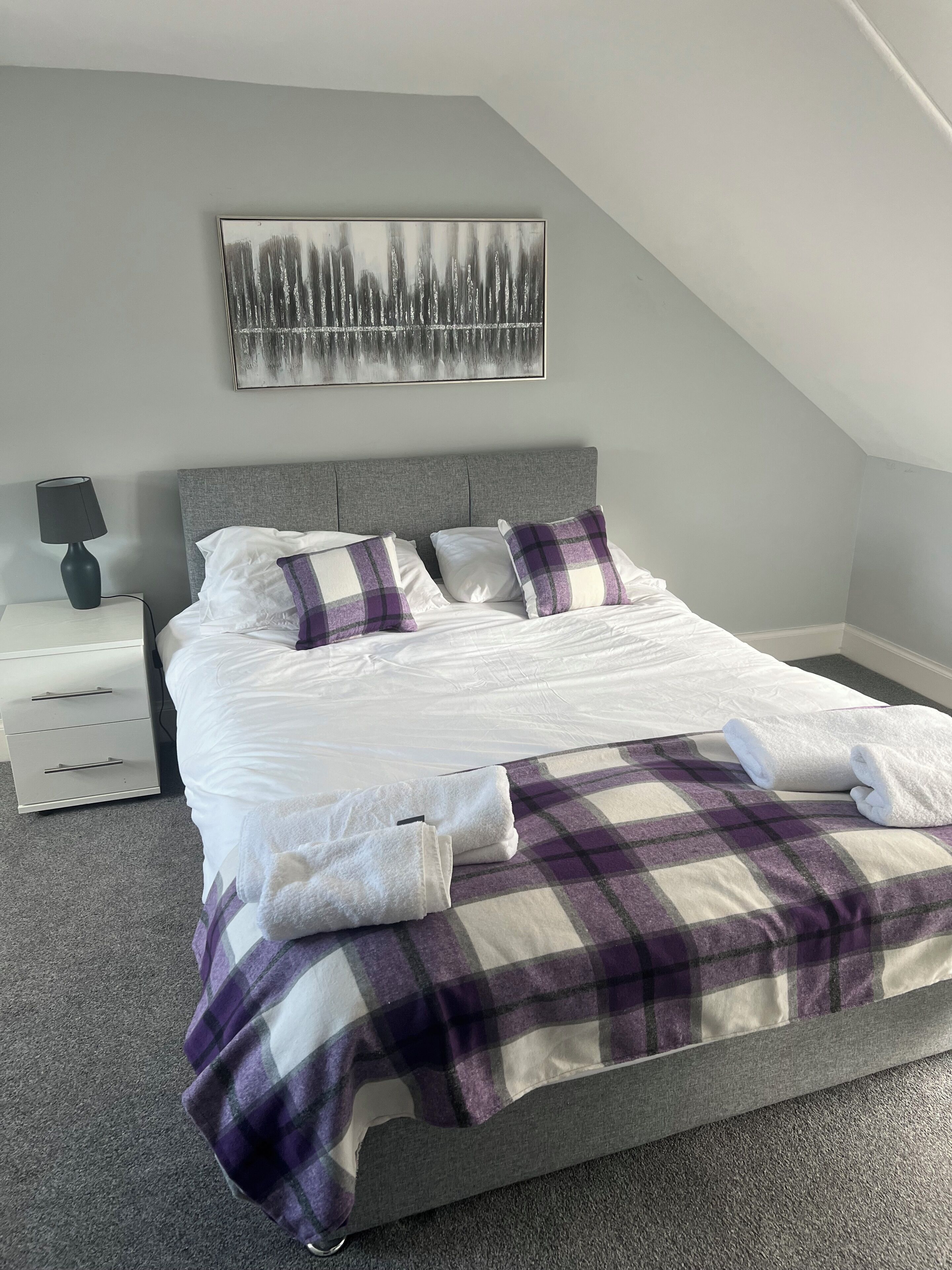 Ein Schlafzimmer mit einem Bett, einem Nachttisch, einer Lampe, einem Gemälde und einer karierten Decke.