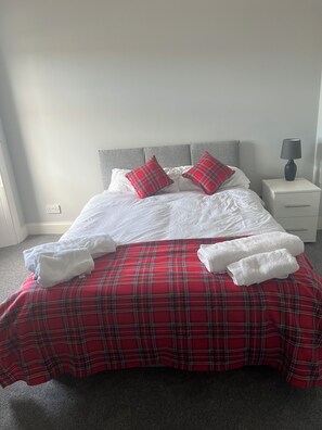 Una cama bien hecha con una colcha escocesa roja y blanca, dos almohadas rojas y blancas, y dos toallas blancas a un lado.