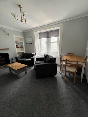 A living room with a fireplace, a sofa, a coffee table, a dining table with chairs, and a window with blinds. -  Our Salad Days Lochgilphead in Lochgilphead town offers a spacious holiday home (Lochgilphead)