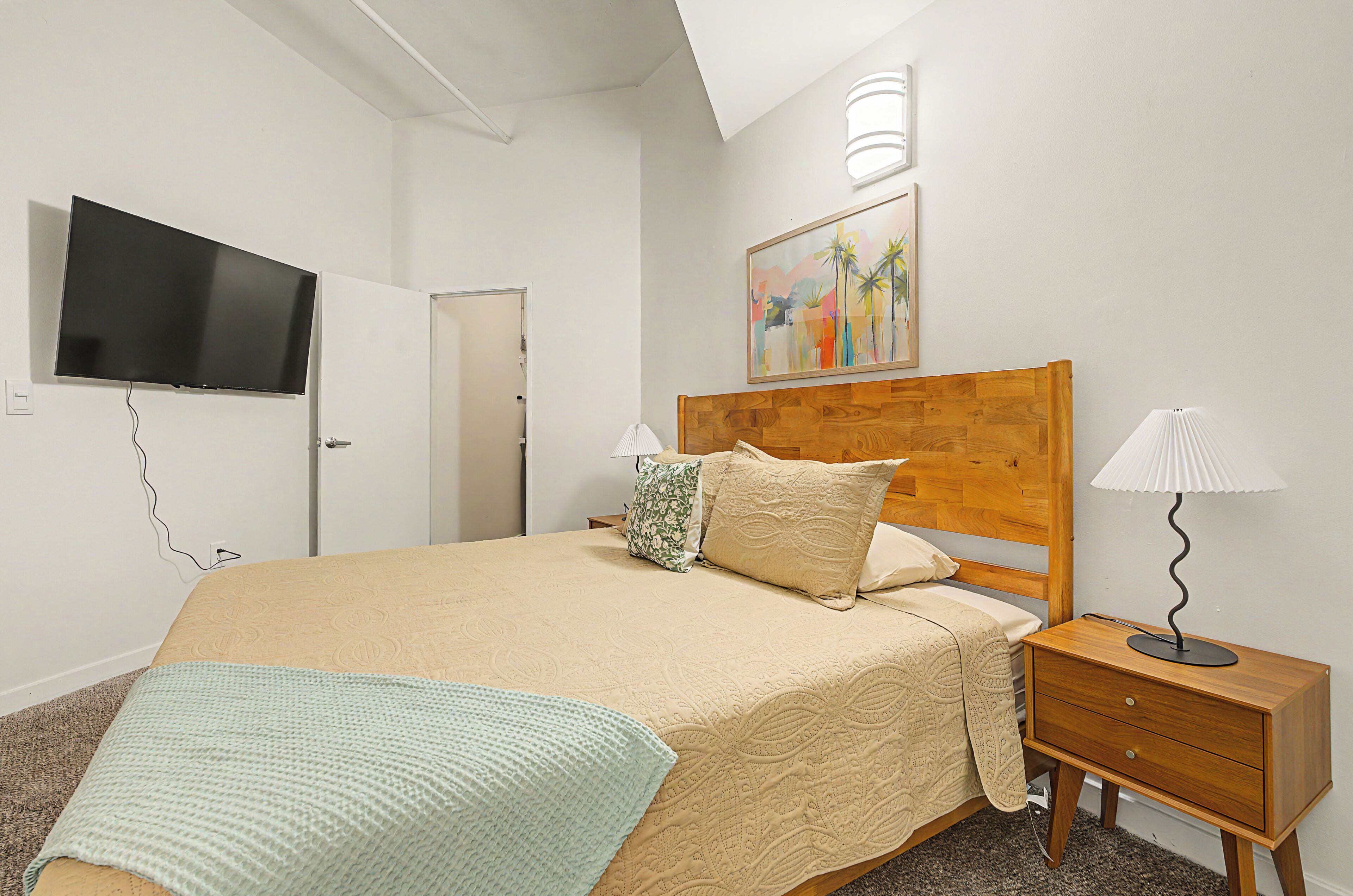 A bedroom with a bed, bedside lamps, a TV, a painting, and a door.