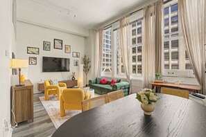 A modern living room with a large flat-screen TV, a dining table, yellow chairs, a green sofa, and a view of a building through large windows. - New Downtown Indy Group Stay +4BR+Sleeps 14 (Indianapolis)
