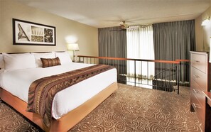 Una habitación de hotel con una cama grande, un ventilador de techo, una mesita de noche con lámpara, un aparador y un cuadro en la pared.
