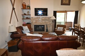 A living room with a leather sofa, a fireplace, a television mounted above it, and a dining area with chairs. - 2 min walk to gondola! (Vail)