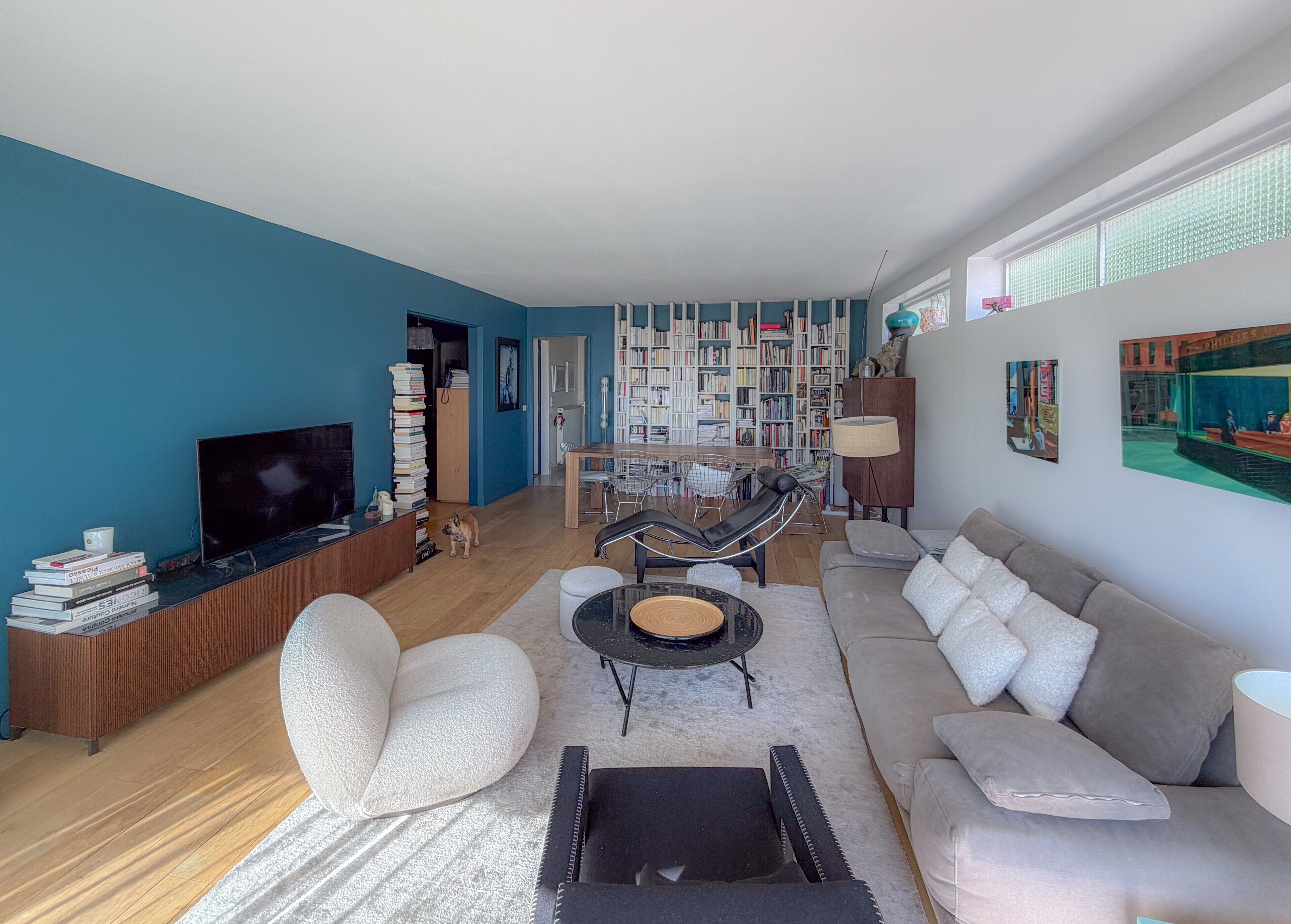 A modern living room with a sofa, armchairs, a coffee table, and a TV. There is a bookshelf filled with books and a dining area with chairs.