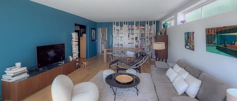 A modern living room with a sofa, armchairs, a coffee table, and a TV. There is a bookshelf filled with books and a dining area with chairs.