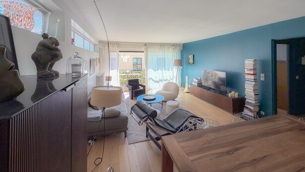A modern living room with a sofa, a coffee table, and a TV. There is a dining table with chairs and a lamp. The room has a large window and a view of the outside. - Beautiful apartment just a stone's throw from Paris, in a quiet neighborhood (Saint-Cloud)