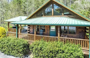 A log cabin with a green metal roof, a porch with seating, and a grill outside. - Lazy Log Cabin at Raccoon Retreat (Whittier)