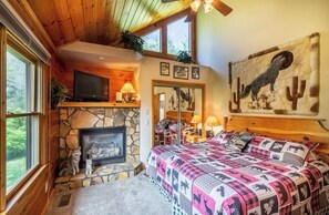 A cozy room with a stone fireplace, a bed with a patterned blanket, and a large painting of a coyote. - Lazy Log Cabin at Raccoon Retreat (Whittier)
