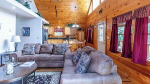 A cozy cabin interior with a wooden ceiling, a living area with a sofa and coffee table, a kitchen with wooden cabinets, and a dining area with a table and chairs. - Lazy Log Cabin at Raccoon Retreat (Whittier)