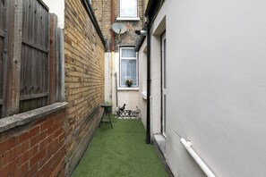 A narrow alleyway with a brick wall on one side, a white wall on the other, and a small patch of artificial grass in the center. - Rose’s Home: Charming 3-Bed House in Stratford (London)