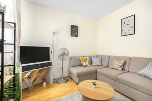 A living room with a flat-screen TV, a standing fan, a sofa with patterned cushions, and a small round coffee table. - Rose’s Home: Charming 3-Bed House in Stratford (London)