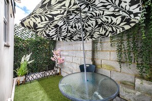 Une table en verre avec un cadre en métal, une chaise, un parapluie à motifs et un jardin agrémenté d’une haie et de fleurs.